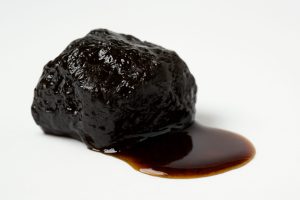 A piece of Shilajit resin with a glossy surface and dark color, showing a small puddle of resin on a white background.